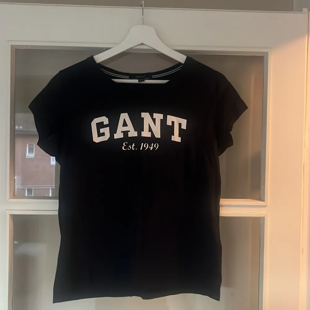 Säljer en snygg svart t-shirt från GANT med vit text 'GANT Est. 1949' på bröstet. Perfekt för en casual look! T-shirten är i mycket bra skick och passar till både jeans och kjol. Skön och stilren design som funkar året runt. . T-shirts.