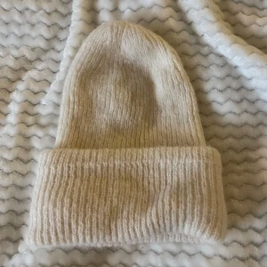 Stickad mössa från Gina Tricot - Säljer en supermysig beige/vit stickad mössa från Gina Tricot. Perfekt för kyliga dagar med sin mjuka och varma känsla. Den har en klassisk design med uppvikt kant som ger en snygg look. Passar perfekt till både vardag och vinteräventyr!