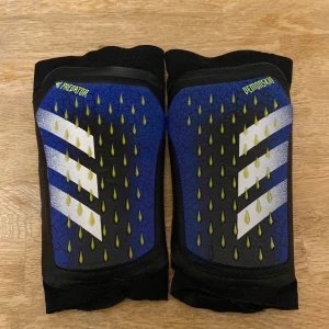 Blå och svarta benskydd från Adidas - Säljer ett par coola Adidas Predator benskydd i blått och svart med gula detaljer. De har en snygg design med vita ränder och är perfekta för fotbollsträningen. Benskydden är märkta med 'Predator' och 'Demonskin' och har en bekväm passform med vaddering på insidan. Passar från 34 till 40.