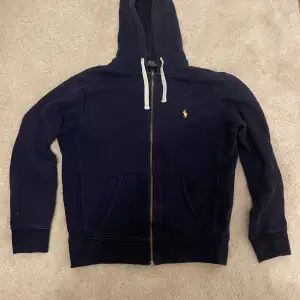 Säljer en snygg mörkblå hoodie från Polo Ralph Lauren i storlek M. Den har en dragkedja framtill och en liten gul logga på bröstet. Perfekt för kyliga dagar och passar till det mesta. Hör av dig vid intresse!