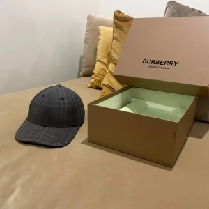 Grå keps från Burberry - Säljer en snygg grå keps från Burberry. Den har en diskret rutig design och är broderad med Burberrys logga baktill. Perfekt för både vardag och lite finare tillfällen. Kepsen är i mycket bra skick och kommer i originalförpackning.
