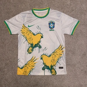 Vit fotbollströja från Nike med fågelmotiv - Säljer en vit Nike fotbollströja med gröna och gula detaljer. Tröjan har ett coolt fågelmotiv i gult och grönt på framsidan och är kortärmad. Perfekt för fotbollsfans som vill sticka ut! 🦜