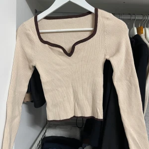 Beige ribbad tröja med brun kant - Säljer en superfin beige ribbad tröja med brun kant runt halsen och ärmsluten. Den är croppad och har en snygg v-ringad design. Perfekt för både höst och vår! Passar till både jeans och kjol för en avslappnad men stilren look.