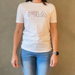 Vit t-shirt från FILA - Säljer en snygg vit t-shirt från FILA i storlek 170-176 cm, perfekt för tonåringar. Den har en klassisk design med FILA-loggan i svart och rött på bröstet. T-shirten är kortärmad och gjord i mjukt bomullsmaterial, vilket gör den superbekväm för vardagsbruk.