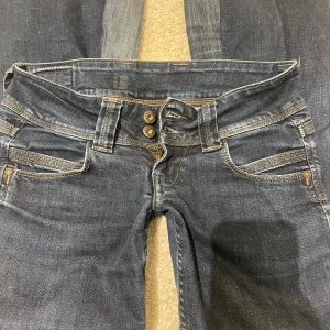 Mörkblå jeans från Pepe Jeans - Säljer ett par snygga mörkblå jeans från Pepe Jeans i bra skick. De har en låg midja och en bootcut-stil. Jeansen har dubbla knappar och dragkedja framtill samt klassiska bakfickor med broderad detalj. Perfekta för både vardag och fest!