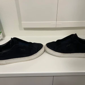 Svarta arigatos utan snören strl:43-44 - Säljer ett par svarta sneakers i mocka med vit sula. De har en stilren design utan snörning, vilket gör dem enkla att ta på och av. Perfekta för vardagsbruk och passar till de flesta outfits. Sulan är lite smutsig men skorna är i bra skick i övrigt. Tillkommer med box och dustbag