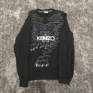 Svart Kenzo tröja med tigertryck - Säljer en svart Kenzo tröja med ett coolt tigertryck i blått och grått på framsidan. Tröjan har långa ärmar och ribbade muddar vid ärmslut och nederkant. Perfekt för en stilren och trendig look. Passar både till vardags och fest!