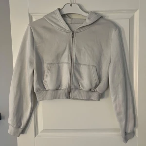 Vit hoodie med dragkedja - Säljer en vit hoodie i bra skick. Den har en dragkedja framtill och en mysig huva. Perfekt för kyliga dagar eller som ett extra lager. Den är croppad och har långa ärmar med ribbade muddar. Passar till både vardag och avslappnade tillfällen.