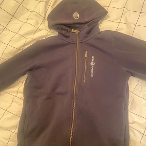 Mörkblå hoodie från Sail Racing - Säljer en snygg mörkblå hoodie från Sail Racing i bra skick. Den har en dragkedja framtill och en mysig huva. Perfekt för höst och vår, och funkar lika bra till vardags som till träning. Den har dock en liten liten defekt. Ens snöret har gått av men det är inget som syns, om den skulle hoppa upp så är det bara och stoppa in den tillbaka i knapphålet. (Den hoppar aldrig upp). Jag som är tjej använder den men har också sätt flera killar ha den. Nypris 1125kr
