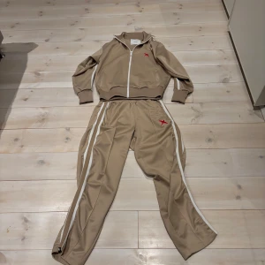 Axel Arigato byxor och tröja - Säljer ett snyggt beige ser från Axel Arigato. Setet består av en jacka med dragkedja och matchande byxor. Båda delarna har vita ränder längs sidorna och en röd logga. Perfekt för en avslappnad stil. Materialet känns mjukt och bekvämt. Man kan köpa byxorna separat och lika så tröjan om man bara är intresserad av något, till ett billigare pris såklart.