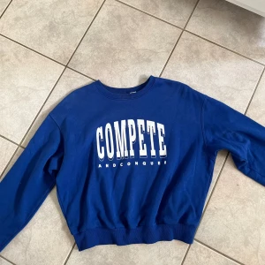 Blå sweatshirt från Divided - Säljer en blå sweatshirt från Divided med texten 'COMPETE AND CONQUER' tryckt på framsidan. Tröjan har en normal passform och är perfekt för en avslappnad stil. Den är i bra skick och redo för nya äventyr! skriv vid frågor💗