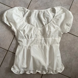 Vit blus med volangdetaljer - brandy Melville liknande! Säljer en söt vit blus med korta puffärmar och volangdetaljer vid nederkanten. Blusen har en v-ringad design med knappar framtill. Perfekt för sommardagar eller en avslappnad vårlook! skriv vid frågor💗