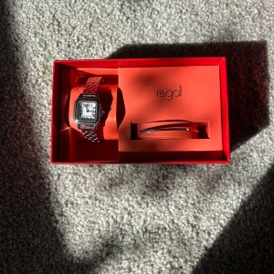 Silverfärgad klocka från Regal - Säljer en stilren och elegant silverfärgad klocka från Regal. Klockan har en fyrkantig urtavla med romerska siffror och ett matchande armband i metall. Perfekt för både vardag och fest! Kommer i en snygg röd presentförpackning.