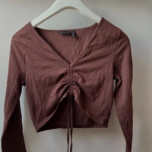 Brun långärmad topp från ASOS DESIGN - Säljer en snygg brun långärmad topp från ASOS DESIGN. Den har en v-ringad design med knytning framtill som ger en cool detalj. Perfekt för höst och vår, och kan stylas både upp och ner beroende på tillfälle.