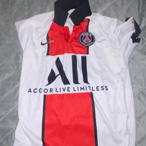 Vit och röd PSG fotbollströja - Säljer en snygg PSG fotbollströja i vitt med röda och svarta detaljer. Tröjan har korta ärmar och är perfekt för alla fotbollsfans. Den har PSG-loggan och Nike-märket på bröstet samt texten 'Accor Live Limitless'.