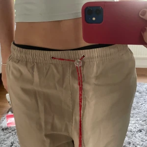 Beige byxor från Le Coq Sportif - Säljer ett par beige byxor från Le Coq Sportif. De har en avslappnad passform med en röd snörning i midjan och röd broderad logga på sidan. Perfekta för hösten. står M men passar även en S beroende på hur baggy man vill ha de