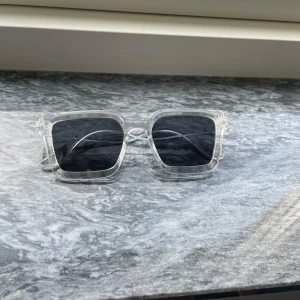 Transparenta solglasögon med fyrkantig form - Snygga och moderna solglasögon med transparent båge och fyrkantiga, mörka linser. Perfekta för en stilren look under soliga dagar. Passar både till vardags och festliga tillfällen. De har en robust design och ger en cool touch till vilken outfit som helst.