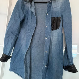 Jeansskjorta från Tingya - Säljer en cool jeansskjorta från Tingya i storlek L. Uppskattar mer som S/M. Skjortan har en unik design med svarta läderdetaljer på fickan och ärmsluten. Perfekt för en avslappnad stil eller en utekväll. Den har knappar framtill. 