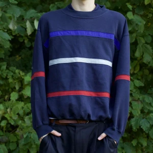 Mörkblå tröja med ränder - Säljer en mörkblå tröja med coola ränder i rött, vitt och blått. Den har en avslappnad passform och långa ärmar, perfekt för höst och vår. Tröjan är i bra skick och passar både till vardags och lite mer avslappnade tillfällen. Skön och stilren, ett måste i garderoben!