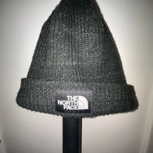 Grå stickad mössa från The North Face - Säljer en snygg och varm grå stickad mössa från The North Face. Perfekt för kyliga dagar med sin ribbade design och uppvikta kant. Den har en klassisk stil som passar till det mesta. Märkesloggan är tydligt broderad på framsidan. Perfekt för både höst och vinter!
