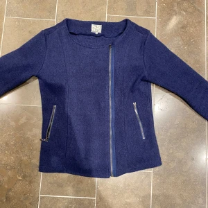 Mörkblå ulljacka med dragkedja - Säljer en snygg mörkblå ulljacka med asymmetrisk dragkedja framtill. Jackan har en stilren design med långa ärmar och två dragkedjefickor. Perfekt för höst och vår när du vill hålla dig varm och stilren. Passar både till vardags och lite finare tillfällen.