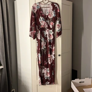 Blommig långklänning från NLY Trend - Säljer en superfin blommig långklänning från NLY Trend i storlek XS. Klänningen har en v-ringad design med trekvartsärm och ett vackert blommönster i rosa och vita nyanser på en vinröd bakgrund. Perfekt för höst och vår, och funkar lika bra till fest som till vardags.