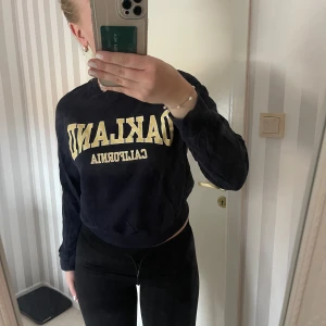 Mörkblå sweatshirt med tryck - Säljer en snygg mörkblå sweatshirt med texten 'OAKLAND CALIFORNIA' i gult på framsidan. Tröjan har långa ärmar och en normal passform, perfekt för en avslappnad stil. Den är i bra skick och passar perfekt till höst och vår.