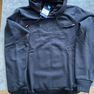 Svart hoodie från GANT - Säljer en snygg och stilren svart hoodie från GANT. Den har en broderad logga på bröstet med texten 'American Sportswear'. Hoodien är i nyskick och har en klassisk passform med långa ärmar och en bekväm huva med dragsko. Stl S tonåring aldrig använd 