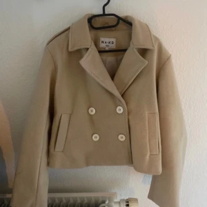 Beige kappa från NA-KD - Säljer en superfin beige kappa från NA-KD i storlek 36. Den är perfekt för höst och vår med sin stilrena design och dubbelknäppning. Jackan är croppad och har långa ärmar, vilket ger en modern look. Den är i nyskick och passar perfekt till både vardag och fest!