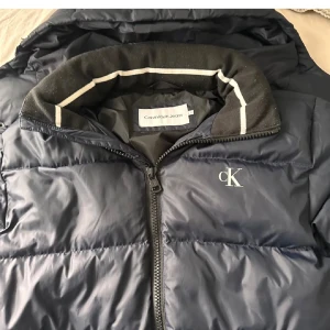 Mörkblå pufferjacka från Calvin Klein Jeans - Säljer en snygg mörkblå pufferjacka från Calvin Klein Jeans. Jackan har en dragkedja framtill och en mysig krage med vita detaljer. Perfekt för kalla dagar med sin vadderade design. Den har också en avtagbar huva för extra skydd mot väder och vind.