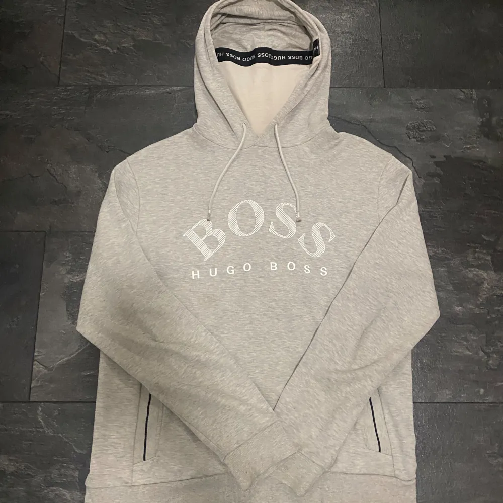 Säljer en snygg och stilren grå hoodie från Hugo Boss. Den har en stor BOSS-logga på framsidan och justerbar dragsko i huvan. Perfekt för en avslappnad och trendig look. Passar både till vardags och chillkvällar.. Hoodies.