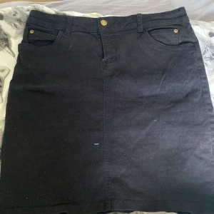 Svart jeanskjol från John Baner - Säljer en snygg svart jeanskjol från John Baner i storlek 40. Kjolen har en klassisk design med fem fickor och en knapp framtill. Perfekt för både vardag och festliga tillfällen. Den är i bra skick och redo för en ny ägare!