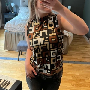 Mönstrad ärmlös topp från Mexx - Säljer en cool ärmlös topp från Mexx med ett unikt geometriskt mönster i svart, brunt och beige. Toppen har en hög krage och en knytning i nacken som ger en snygg detalj. Axelbredd: 38 cm och Längd:  62 cm.