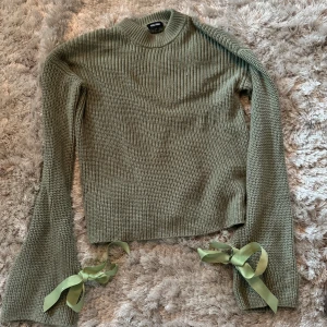 Grön stickad tröja med rosetter - Säljer en superfin grön stickad tröja med långa ärmar och söta rosetter vid ärmsluten. Perfekt för höst och vinter, den är både mysig och stilren. Passar perfekt till både jeans och kjol!