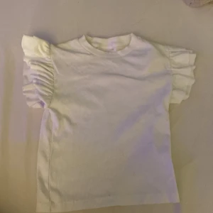 Kort vit ribbad tshirt/top med volang i storlek 146/152 - Jättefin kort vit ribbad tshirt/top med volang i storlek 146/152, köpt från Gina tricot och jag säljer pga inte min stil❤️