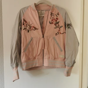 Rosa jacka med broderier från Odd Molly - Säljer en superfin rosa jacka från Odd Molly med vackra broderier av blommor och fjärilar. Jackan har en dragkedja framtill och grå ärmar som ger en snygg kontrast. Perfekt för vår och sommar! 🌸