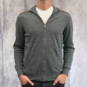 Grå tröja med dragkedja från Massimo Dutti - Säljer en stilren grå tröja från Massimo Dutti i storlek M. Tröjan är tillverkad i en mjuk kashmirblandning och har en dragkedja framtill. Den har långa ärmar och ribbade muddar, vilket gör den perfekt för både höst och vinter. Passar både till vardags och lite finare tillfällen.