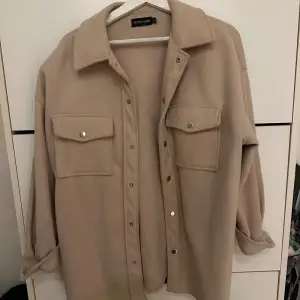 Säljer en snygg beige overshirt från Bershka. Den har en avslappnad passform med långa ärmar och knappar framtill. Två stora fickor på bröstet ger en cool look. Perfekt för lager-på-lager under hösten eller våren! Använd 1 gång.