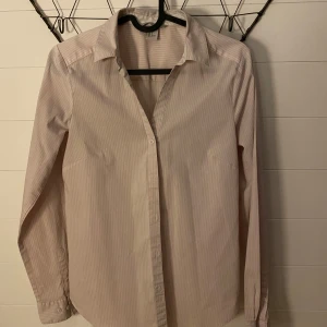 Randig skjorta med långa ärmar - Säljer en stilren och klassisk randig skjorta med långa ärmar. Skjortan har en krage och knappar framtill. Perfekt för både vardag och festliga tillfällen!