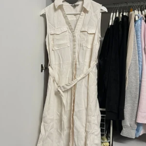 Vit ärmlös klänning från H&M - Säljer en stilren vit ärmlös klänning från H&M i storlek M. Klänningen har en klassisk krage och två bröstfickor som ger en avslappnad look. Den har ett bälte i midjan för att framhäva figuren. Perfekt för sommarens alla tillfällen! Aldrig använd