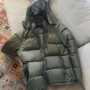 Grön pufferjacka med huva - Säljer en snygg och varm grön jacka  från Ralph Lauren. Jackan har en praktisk huva och dragkedja. Perfekt för kalla dagar och håller dig varm och stilren. Den är i bra skick, men har upptäckt små defekter på insidan av jackan som ej märkbara. 