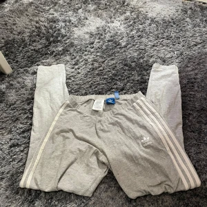 Grå mjukisbyxor från Adidas - Säljer ett par grå mjukisbyxor från Adidas. De har de klassiska tre ränderna längs sidorna och en elastisk midja för extra komfort. Perfekta för en avslappnad dag hemma eller lätt träning. Byxorna är i bra skick och redo för en ny ägare!