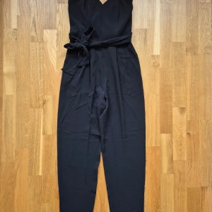 Byxdress HM - Svart byxdress från HM. Nyskick, aldrig använd. Bälte runt midjan. Korsett lik överdel. Byxfickor finns på vardera sida. Dold dragkedja på ena sidan. Resårband på ryggen. Storlek 34. 96% polyester, 4% elastan.