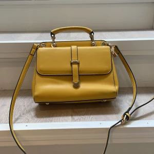 Gul axelväska med handtag - Säljer en superfin gul axelväska med ett praktiskt handtag. Väskan har en stilren design med en justerbar axelrem och ett snyggt spänne framtill. Perfekt för både vardag och fest!