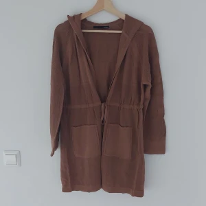 Brun finstickad kofta från LeComte - Säljer en mysig brun  finstickad kofta från LeComte. Den har en huva och knytband i midjan för en bekväm passform. Perfekt för höst och vinter, med långa ärmar och praktiska fickor framtill. Superfin att ha på sig nu i höst!