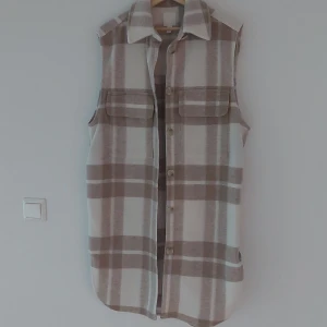 Rutig väst från H&M - Säljer en superfin rutig väst från H&M i storlek M. Västen har en beige och vit färgkombination med stora rutor och knappar framtill. Den har en klassisk krage och två stora fickor på bröstet. Perfekt för höst och vår!