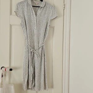 Vit prickig klänning med knytband - Säljer en superfin vit klänning med svarta prickar. Den har korta ärmar och ett knytband i midjan för en snygg passform. Perfekt för sommarens alla fester eller en avslappnad dag på stan. Klänningen är i nyskick och redo för nya äventyr!