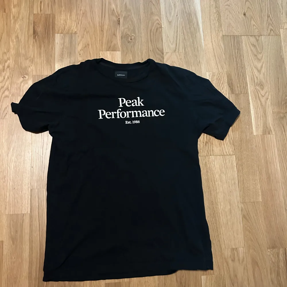 Säljer en svart t-shirt från Peak Performance med vit text på framsidan. Den är i bra skick och perfekt för vardagsbruk. Storleken är 170, vilket motsvarar ungefär en herr M. T-shirten är gjord i mjuk bomull och har en normal passform. Perfekt för både träning och avslappnade dagar!. T-shirts.