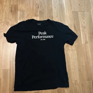 Svart t-shirt från Peak Performance - Säljer en svart t-shirt från Peak Performance med vit text på framsidan. Den är i bra skick och perfekt för vardagsbruk. Storleken är 170, vilket motsvarar ungefär en herr M. T-shirten är gjord i mjuk bomull och har en normal passform. Perfekt för både träning och avslappnade dagar!