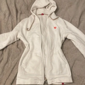 Vit fleecejacka med huva - Mysig vit fleecejacka med huva och dragkedja. Perfekt för kyliga dagar. Jackan har två fickor framtill och en liten röd logga på bröstet. Den är långärmad och har en avslappnad passform.